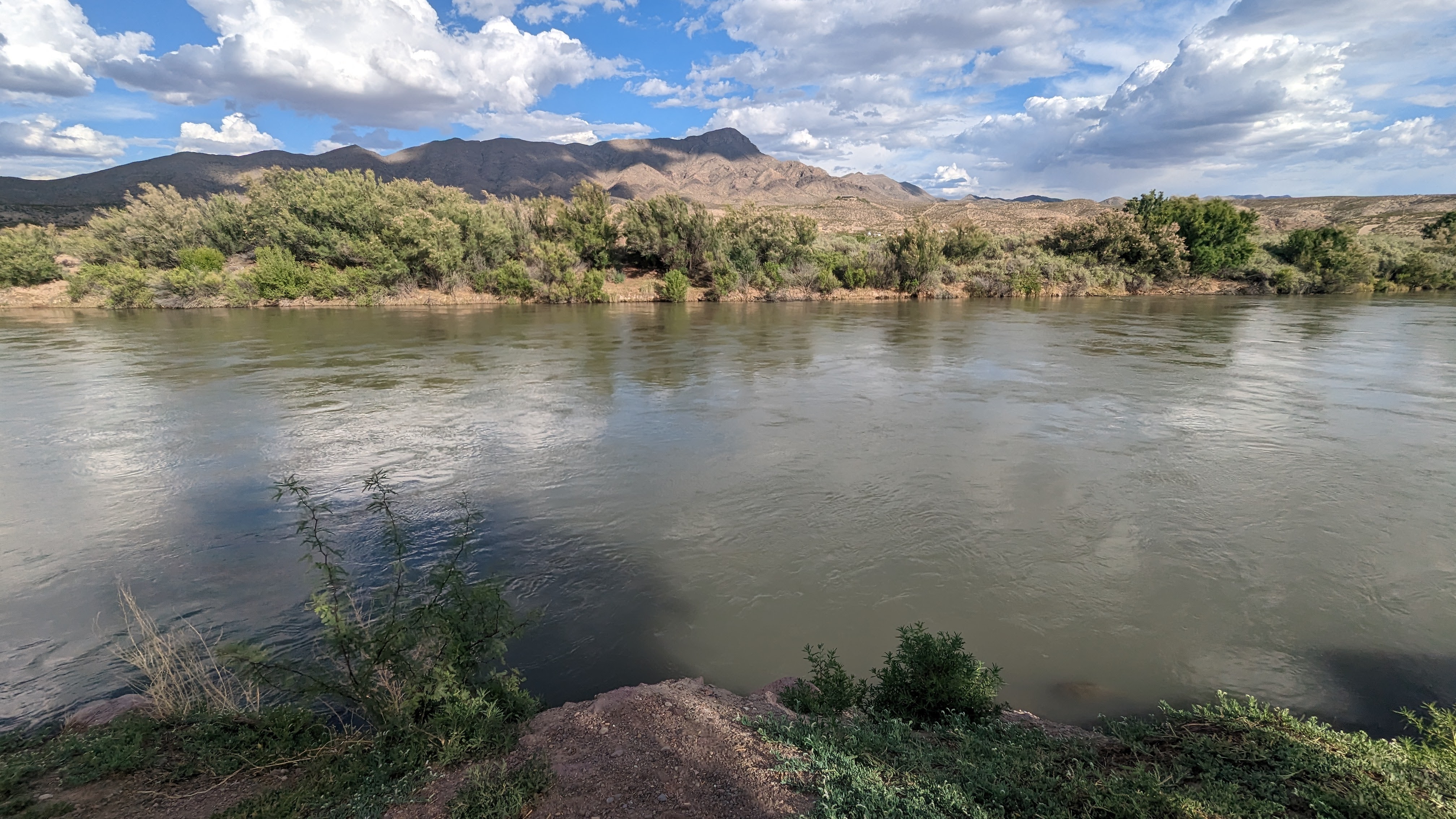 The Rio Grande in May, Truth or Consequences, New Mexico. Mountains in the distance. Blue sky with clouds, murky water. Green ground cover over brown sand.