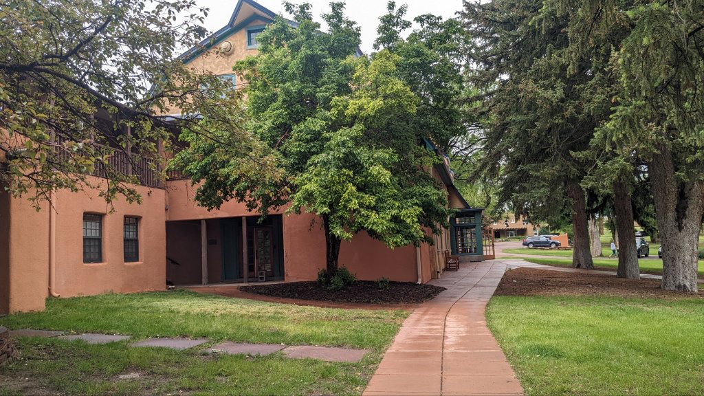 Path leading to the arts building at Fountain Valley School. Red concrete, adobe buildings, tall green trees, and grass.