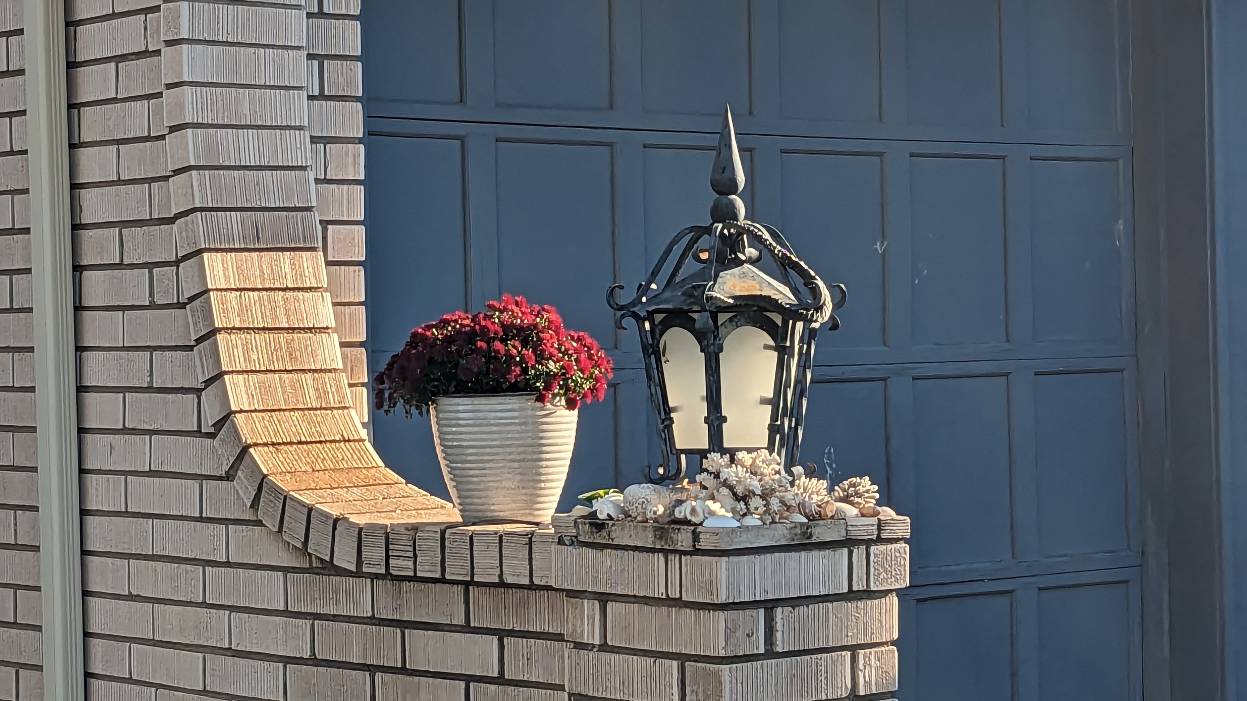 Outdoor light and a plant with red flowers sitting on a brick half-wall.
